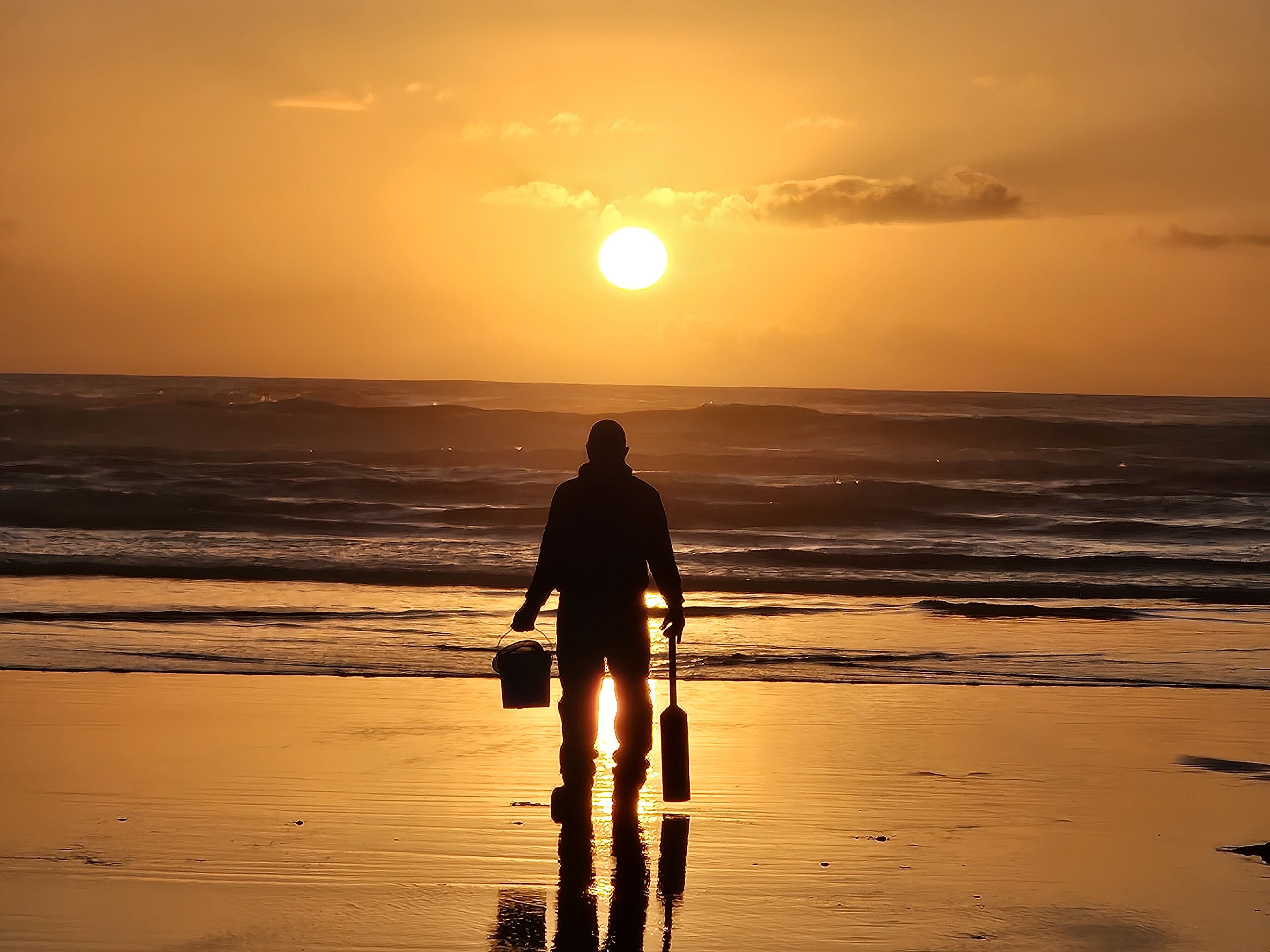 Person holding a small bucket and clam gun on the beach at sunset.