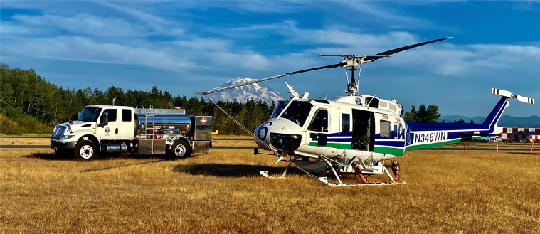 A helicopter and a fuel truck are parked on a grassy field, with a mountain in the background under a clear blue sky.