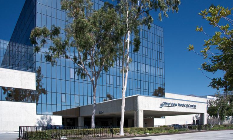 A modern medical center building with large glass windows, surrounded by trees and a clear blue sky.