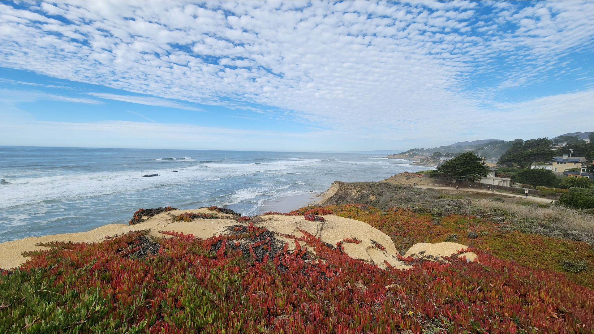 A coastal view featuring waves, sandy cliffs, and colorful vegetation under a partly cloudy sky.