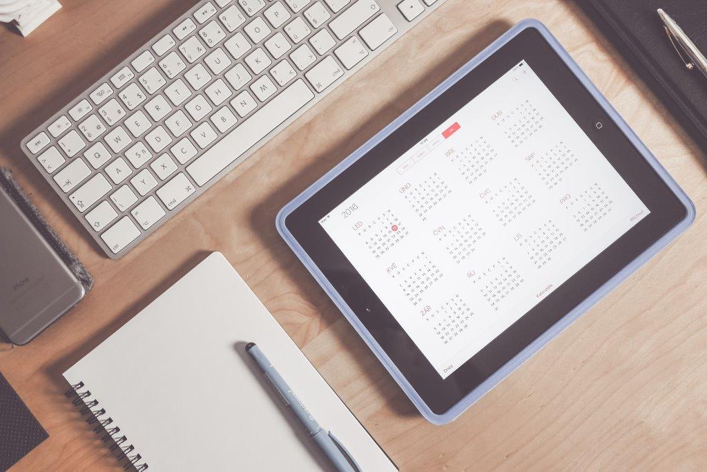 A workspace featuring a tablet displaying a calendar, a keyboard, a notebook, and a pen on a wooden surface.